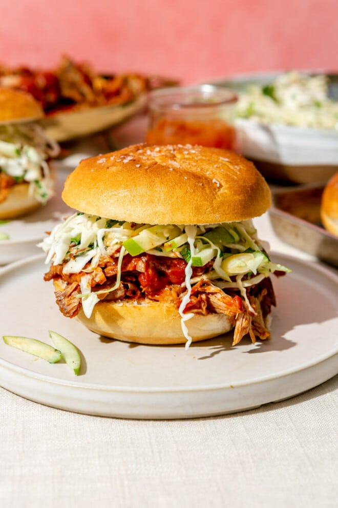 A pulled pork sandwich topped with green apple slaw in front of a large bowl of slaw, a large bowl of pulled pork, and another pulled pork sandwich.