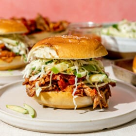 A pulled pork sandwich topped with green apple slaw in front of a large bowl of slaw, a large bowl of pulled pork, and another pulled pork sandwich.