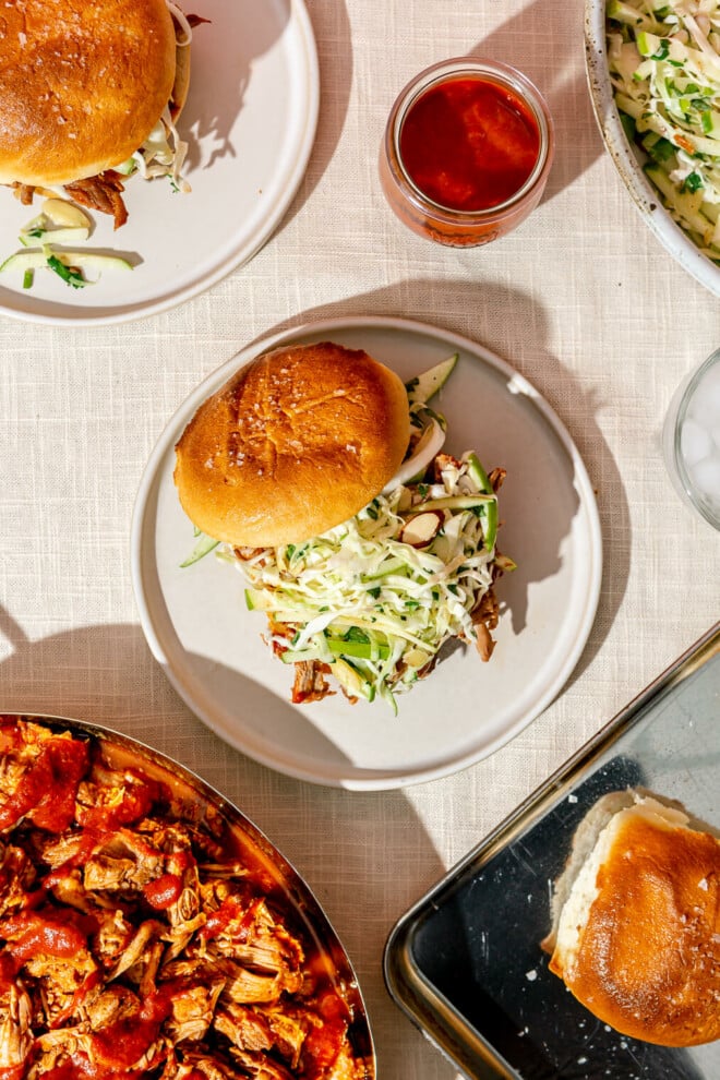 A pulled pork sandwich topped with green apple slaw in front of a large bowl of slaw, a large bowl of pulled pork, and another pulled pork sandwich.