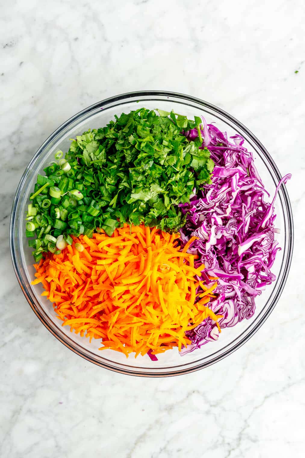 A top down view of a large glass bowl with purple cabbage, shredded carrots, cilantro, and green onions.