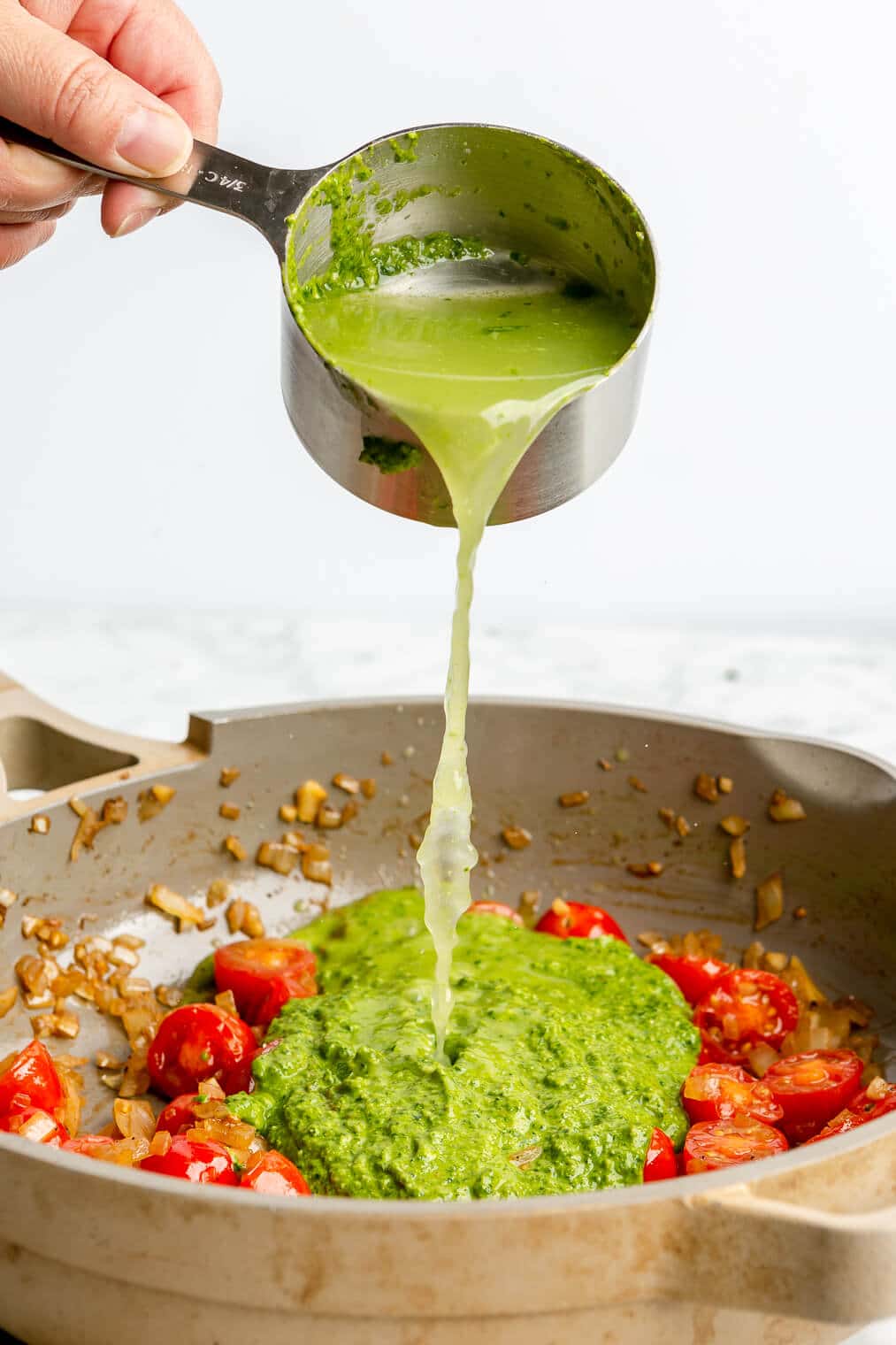 A person adding broth to a pan of sauteed onions, cherry tomatoes, and pesto sauce.