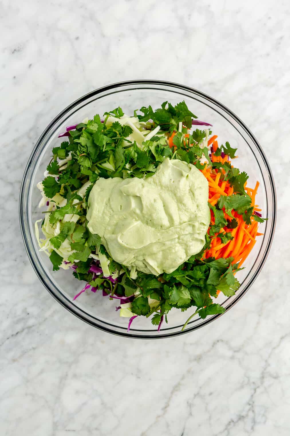 Slaw ingredients in a large glass bowl topped with avocado cream sauce on a grey and white marble surface.