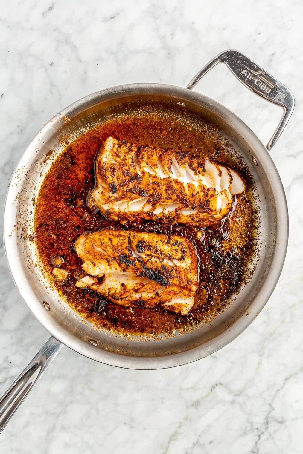 Cod filets coated in blackened seasoning in a large skillet with butter that has been cooked and seared on one side.