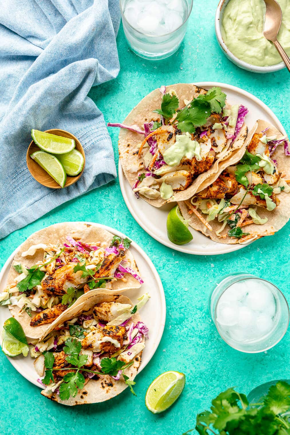 Two plates of blackened fish tacos on round plates garnished with cilantro and a lime wedge. The plates are on a teal surface with glasses of ice water and a light blue linen draped in the top right corner. There is a small, wooden serving bowl with lime wedges and a white bowl with avocado cream with a bronze spoon.