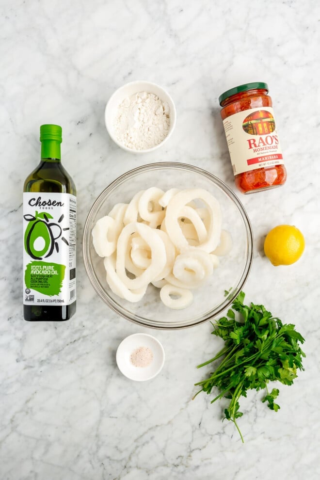 All of the ingredients for fried calamari (squid, avocado oil, flour, lemon, parsley, salt, and marinara sauce) sitting on a marble surface.
