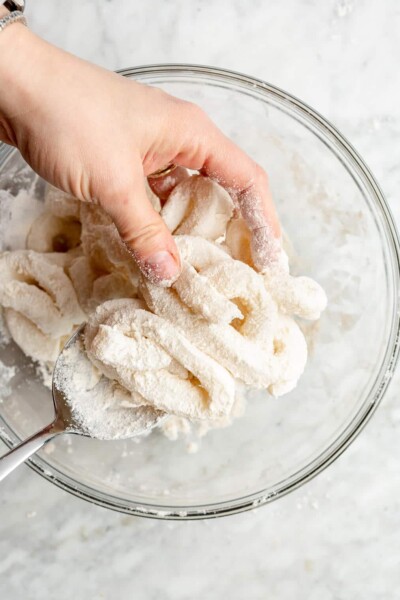 A person using their hand and a spoon to toss calamari rings in a bowl of flour and salt.