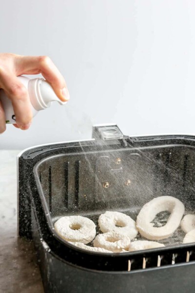 A person spraying oil onto raw, breaded calamari rings in an air fryer basket.