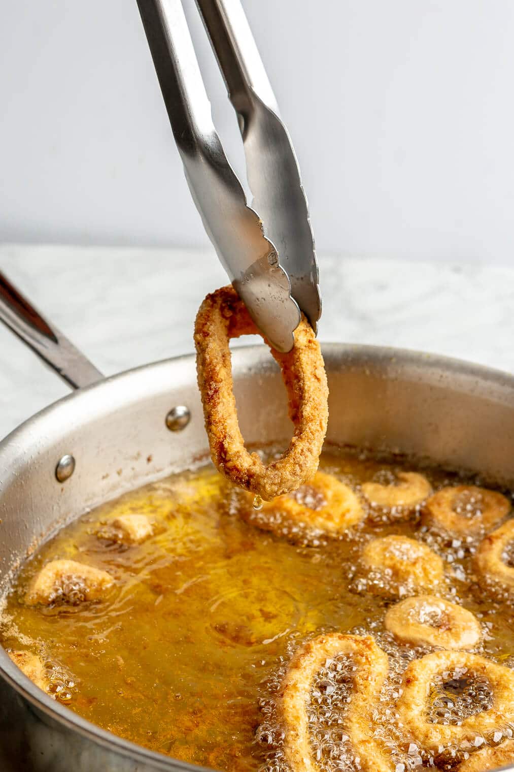 A person using metal tongs to take a fried calamari ring out of a pan of hot oil while more calamari rings continue to fry.