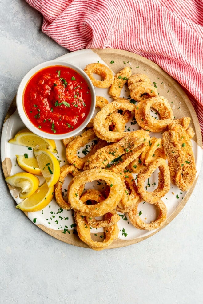 Top view of a plate of crispy fried calamari rings next to lemon wedges and a small bowl of marinara sauce.