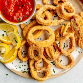 A top view of crispy fried calamari on a plate next to lemon wedges and a bowl of marinara.