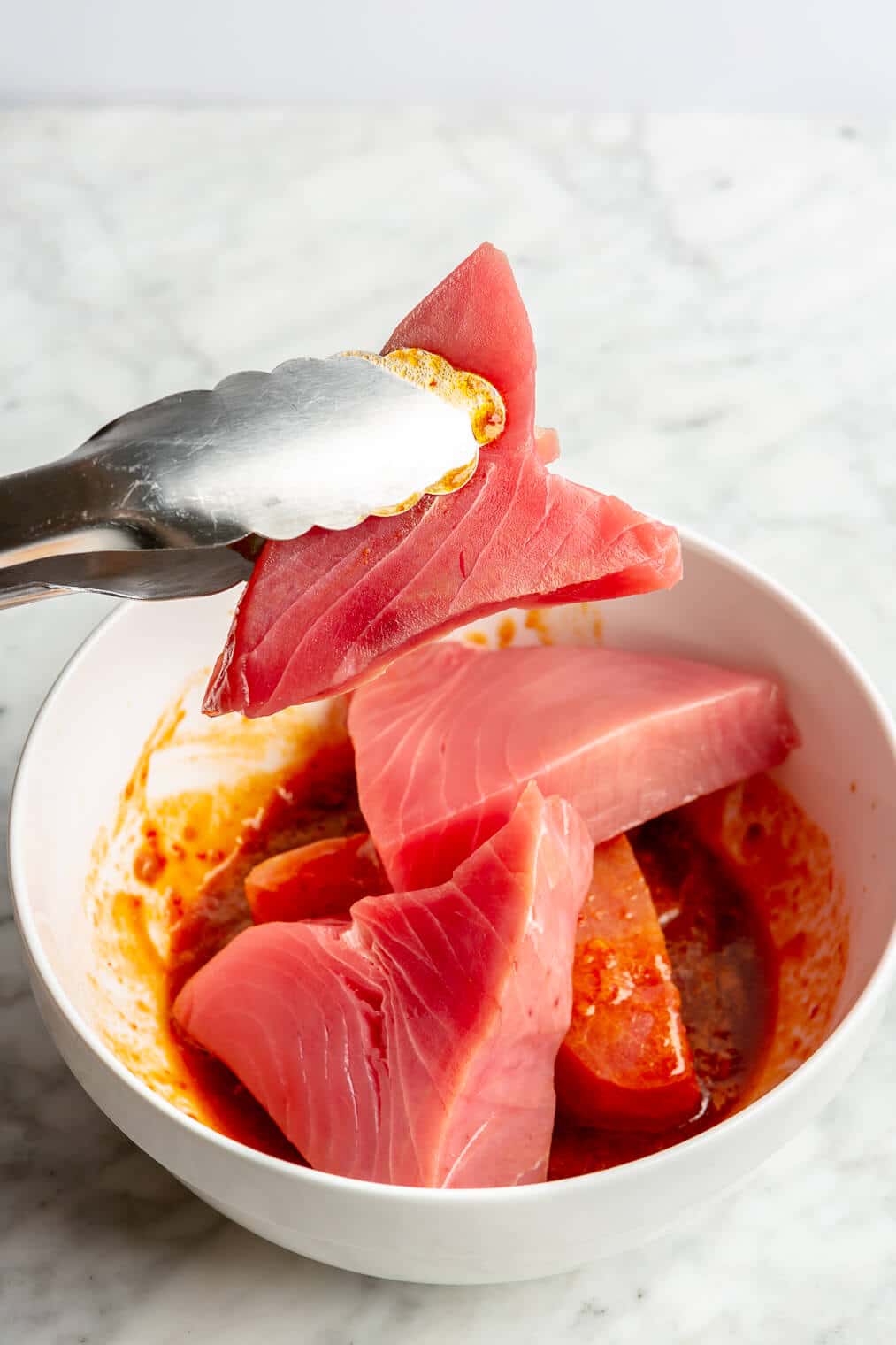 A pair of tongs holding up a tuna steak over a white bowl with chili-lime marinade.
