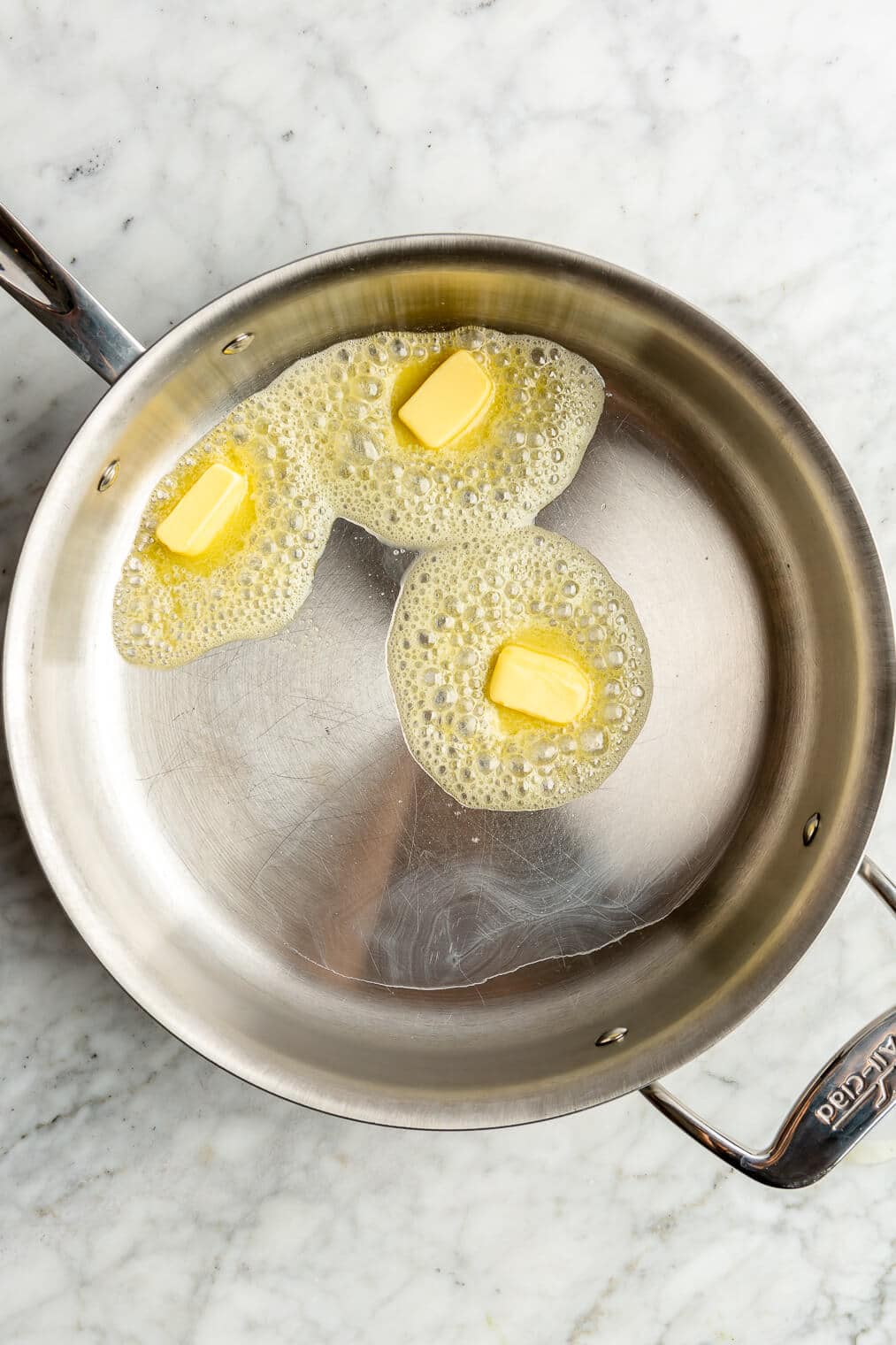 Butter melting and bubbling in an all-clad pan on a grey and white marble surface.