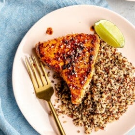 Chili lime seared tuna steak on a light pink plate with a gold fork and side of quinoa with a lime wedge. The plate is on a light grey surface with a light blue linen. There is another plate to the top with a serving of quinoa and a small bowl of chili sauce on the bottom right.
