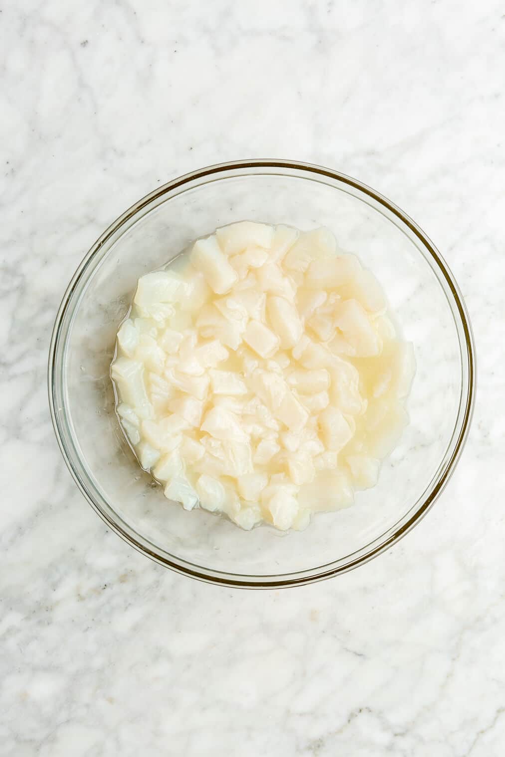 Bowl of diced white fish with lime juice in a large glass bowl on a grey and white marble surface.