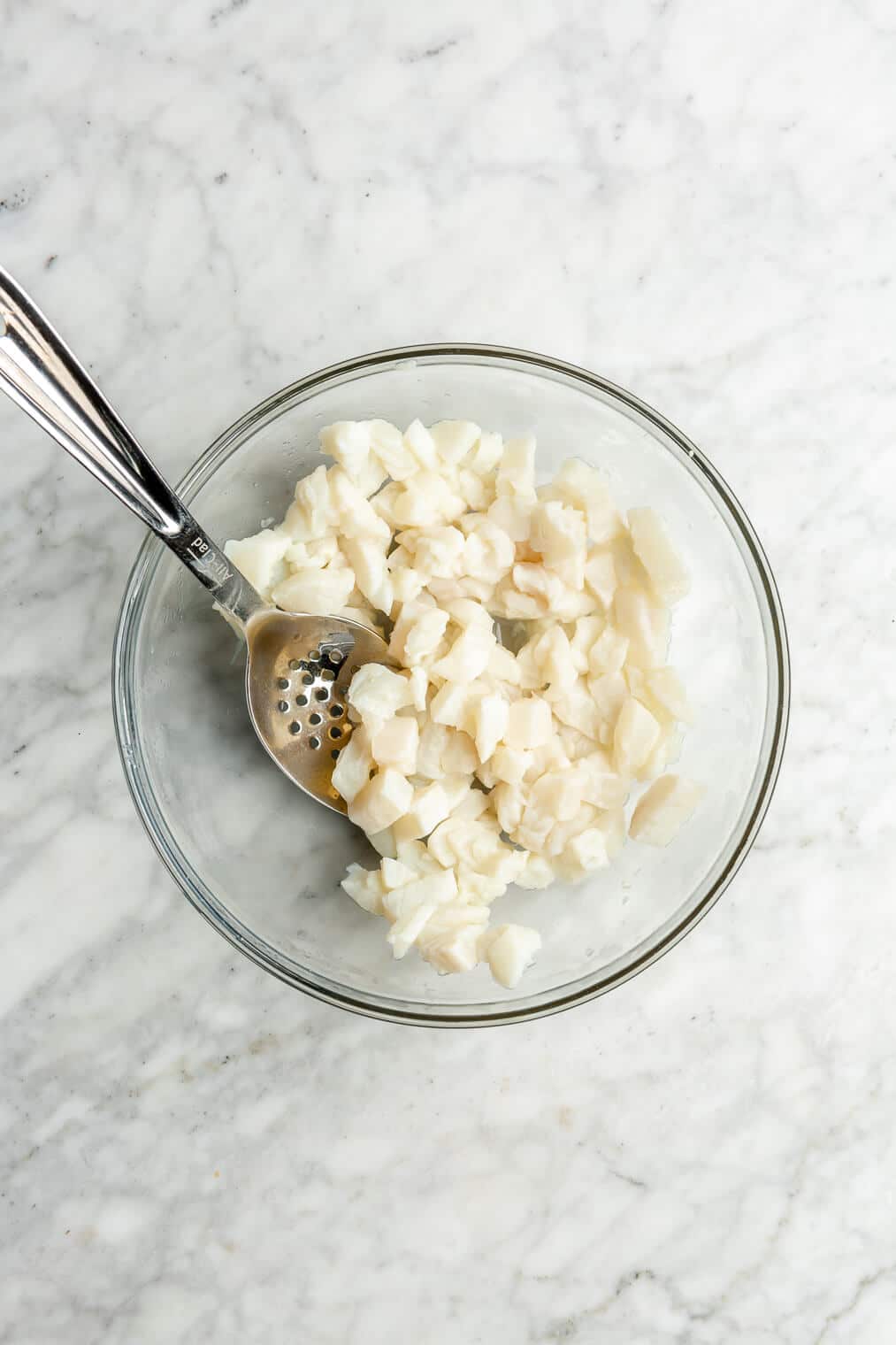 Large glass bowl with marinated white fish cut into cubes. There is an all-clad stainless steel slotted spoon resting in the bowl.