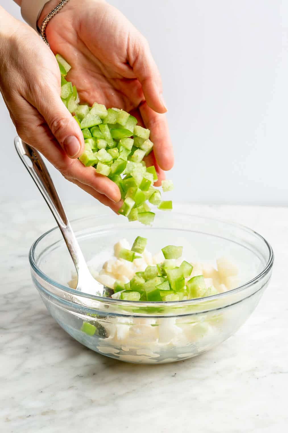 Hands dropping diced tomatillos into a large glass bowl with cubed white fish.