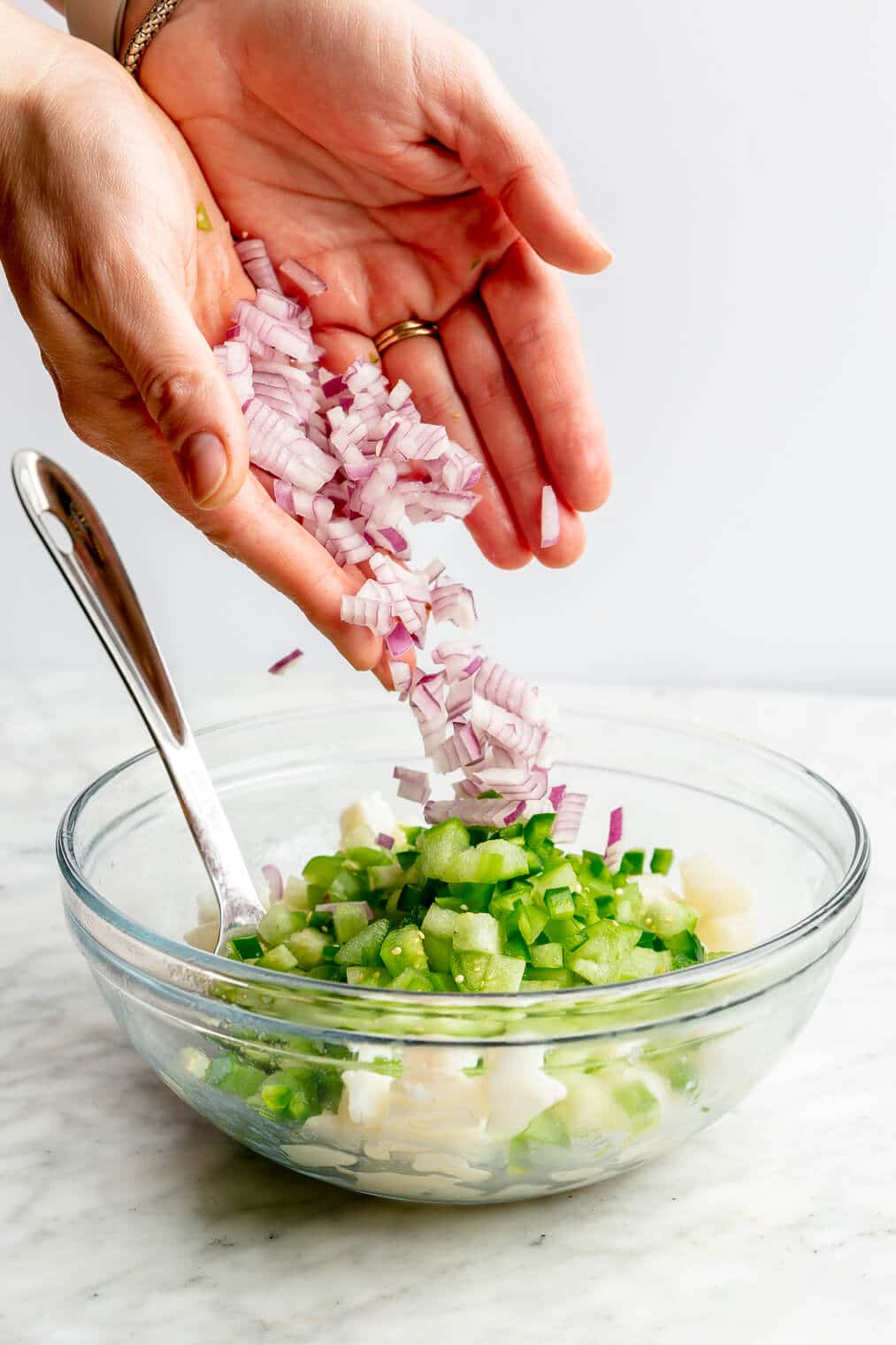 Hands dropping diced red onion into a large glass bowl with cubed white fish and veggies.