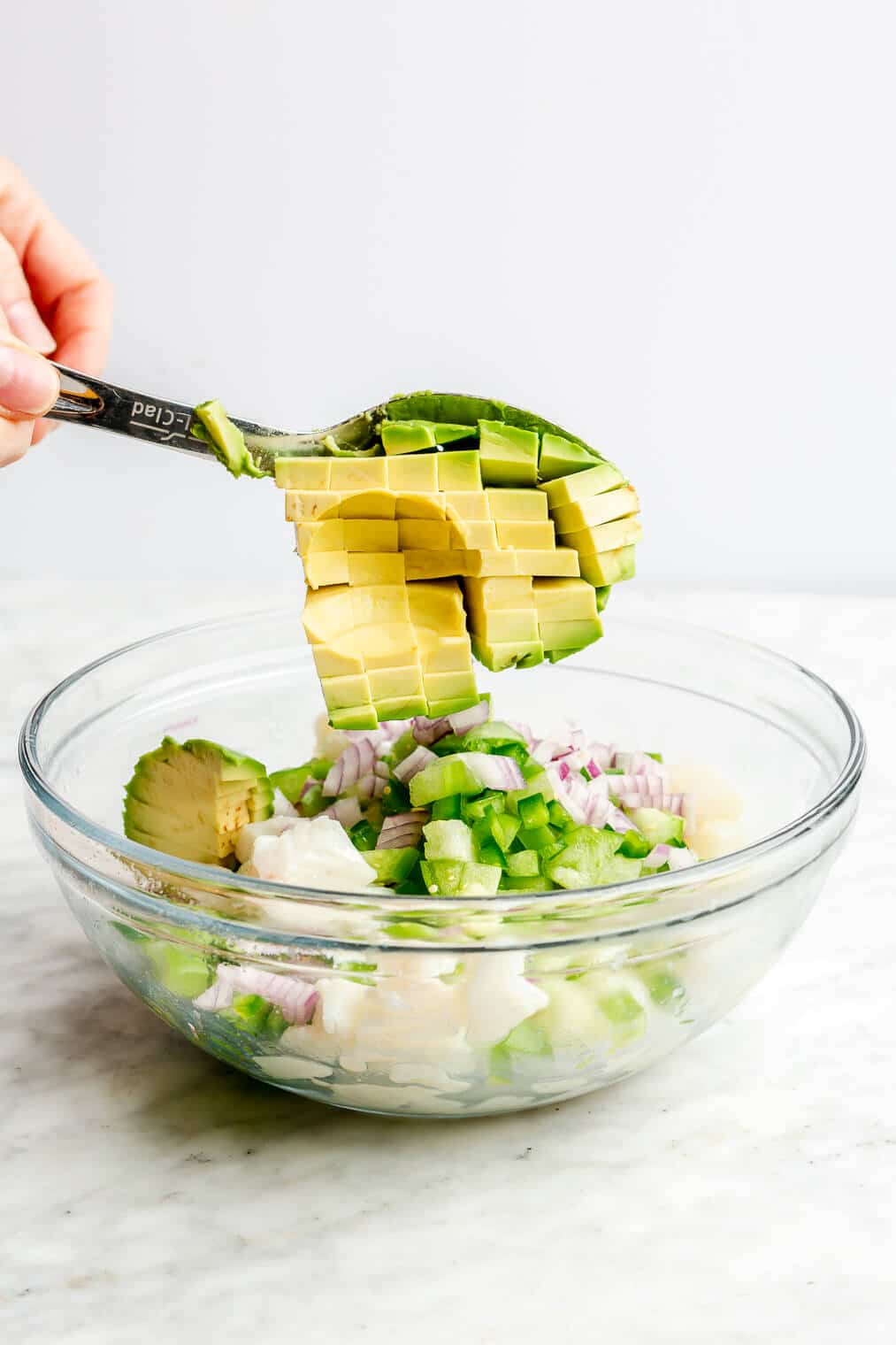 Hand spooning diced avocado into a large glass bowl with cubed white fish and veggies.