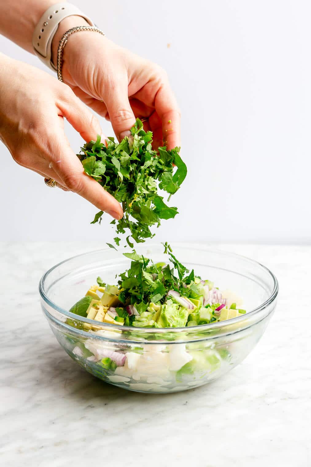 Hand dropping chopped cilantro into a large glass bowl with cubed white fish and veggies.