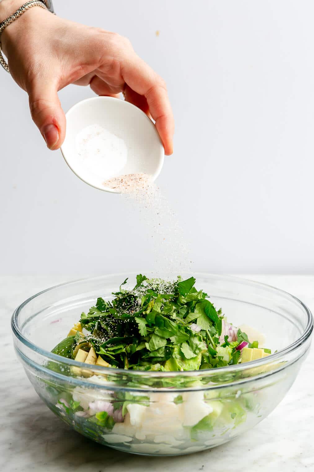 Hand pouring salt over top of cilantro and diced veggies with cubed white fish in a large glass bowl.