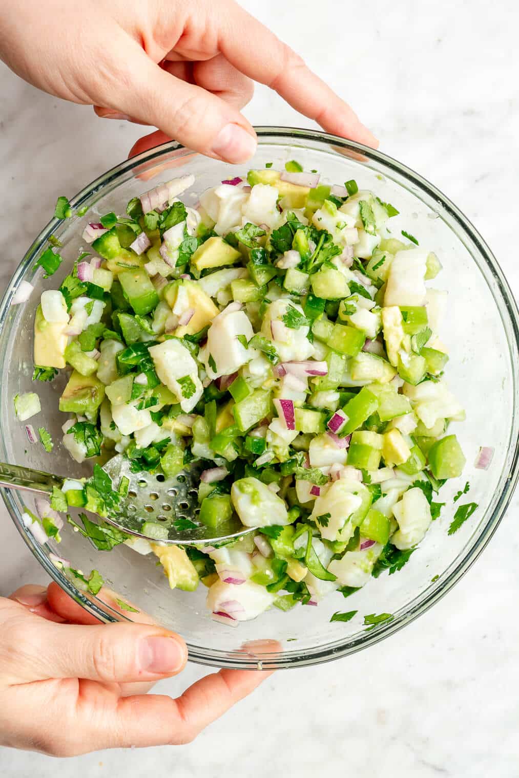 Hands holding a large glass bowl with white fish ceviche mixed with diced tomatillos, avocado, jalapeno, and cilantro.