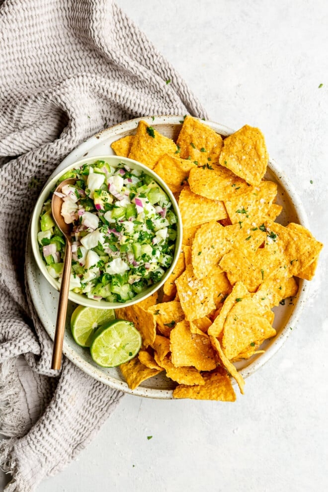 A large, shallow bowl with tortilla chips and a bowl of fish ceviche verde in a white bowl on the side with a bronze spoon. The plate is garnished with two lime halves and some flaky sea salt. There is a light grey, textured linen draped to the left side.