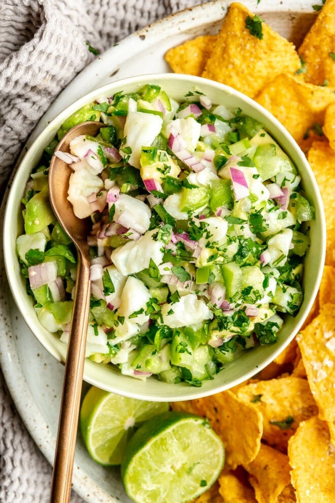 Bowl of fish ceviche verde with a bronze spoon on a plate with tortilla chips garnished with a couple of lime halves and flaky sea salt. There is a textured grey linen to the side of the plate.