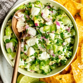 Bowl of fish ceviche verde with a bronze spoon on a plate with tortilla chips garnished with a couple of lime halves and flaky sea salt. There is a textured grey linen to the side of the plate.