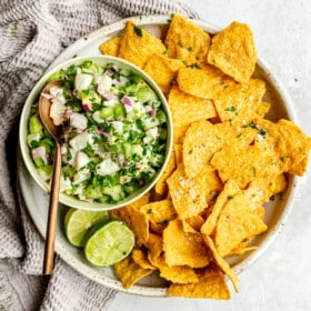 A large, shallow bowl with tortilla chips and a bowl of fish ceviche verde in a white bowl on the side with a bronze spoon. The plate is garnished with two lime halves and some flaky sea salt. There is a light grey, textured linen draped to the left side.