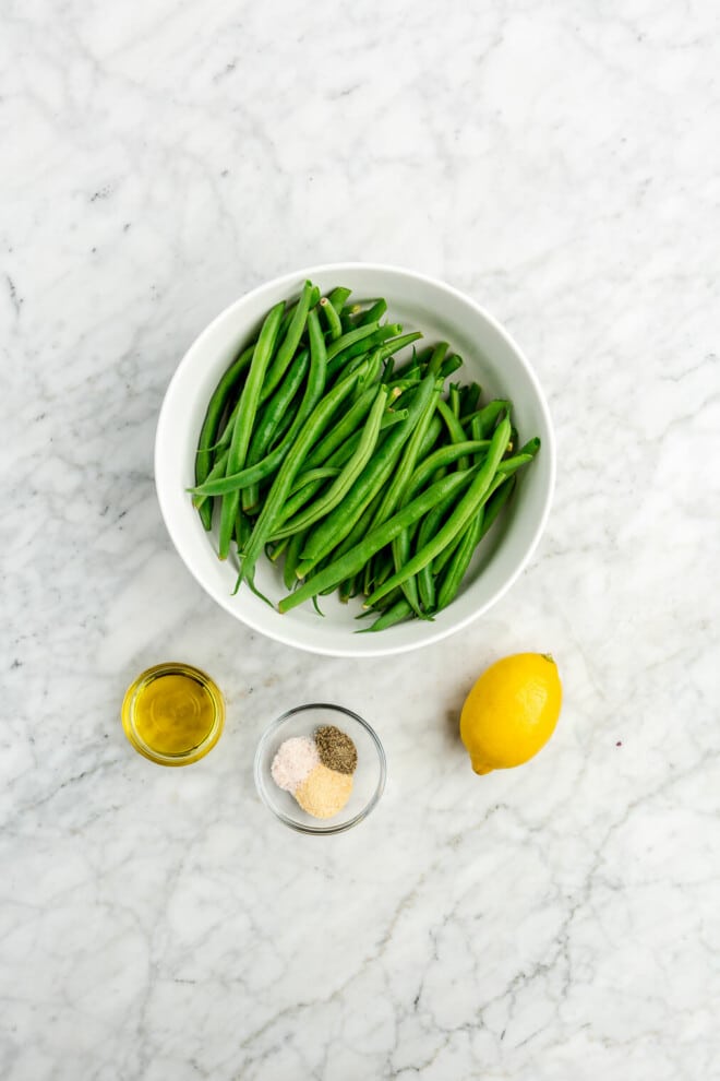 All of the ingredients needed for oven roasted green beans (fresh green beans, olive oil, salt, pepper, and lemon) on a marble surface.