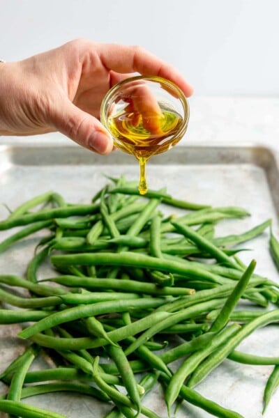 A person pouring a small bowl of olive oil onto a sheet pan of fresh green beans.