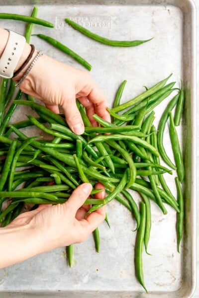 A person using their hands to toss fresh green beans with olive oil on a sheet pan.