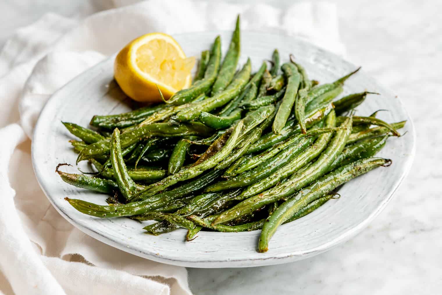 A white bowl filled with oven roasted green beans and garnished with a half squeezed lemon. There is a white linen draped to the left side.