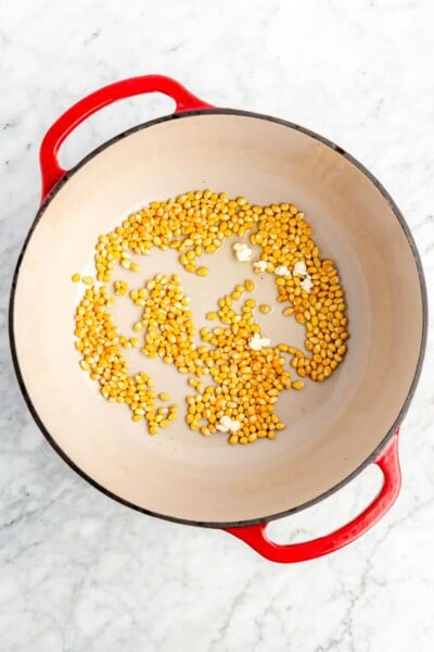 A red dutch oven with a white enamel interior with olive oil and corn kernels in the oven.
