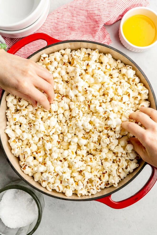 A red dutch oven with white interior with popped corn on a grey surface. There are two hands reaching for the popcorn in the pot. There is a glass jar of flaky sea salt, a white bowl with melted butter, and a red and white striped linen on the table.