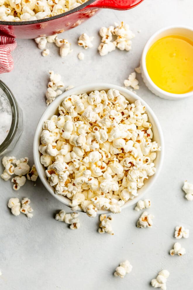 A white bowl of popped corn overflowing on a grey surface. There is a white bowl of melted butter and a glass jar with flaky sea salt on the surface.