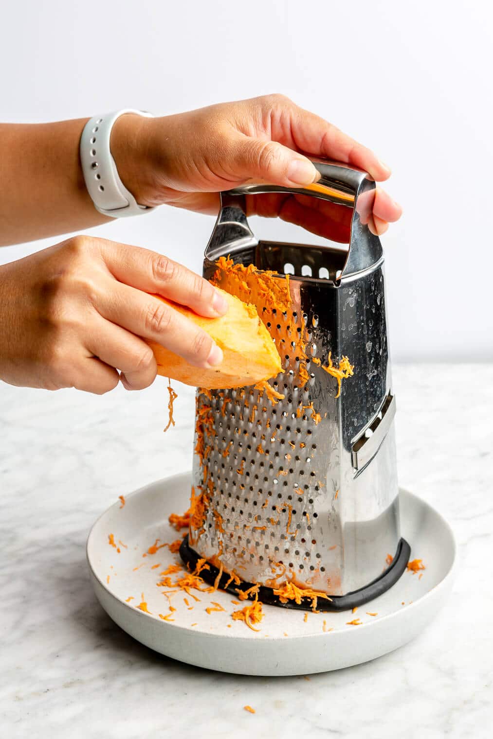 Hand grating a sweet potato using a hand grater over a plate on a grey and white marble surface.