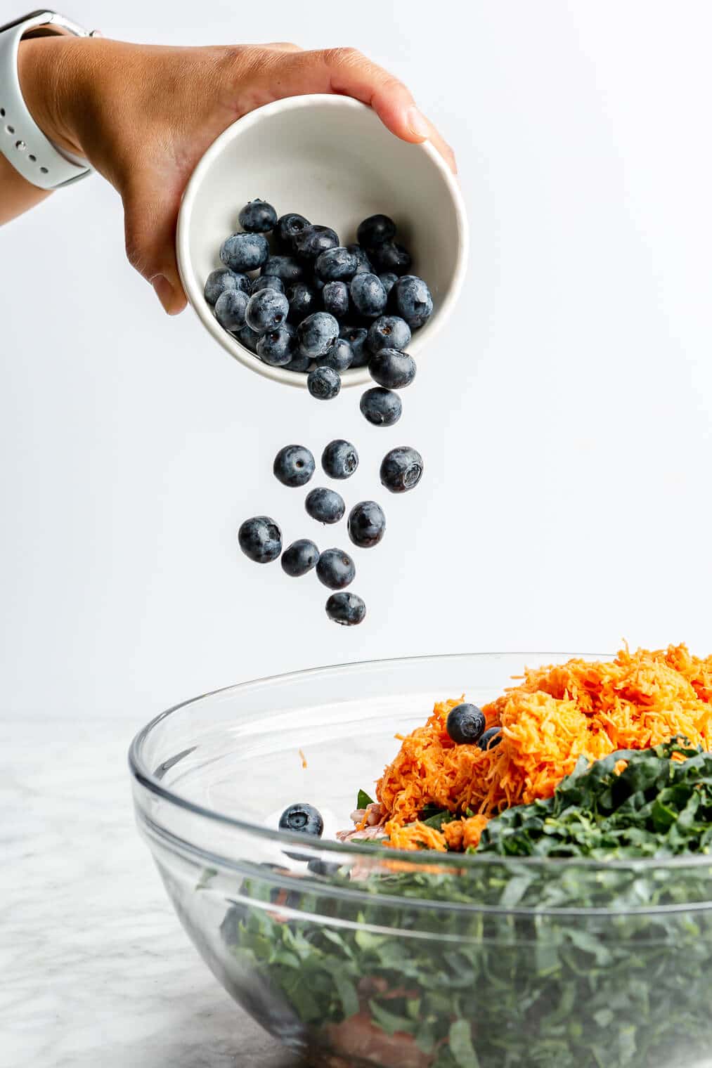 Hand pouring blueberries out of a small, white bowl into a large glass bowl with ground pork, shredded sweet potato, and shredded kale.