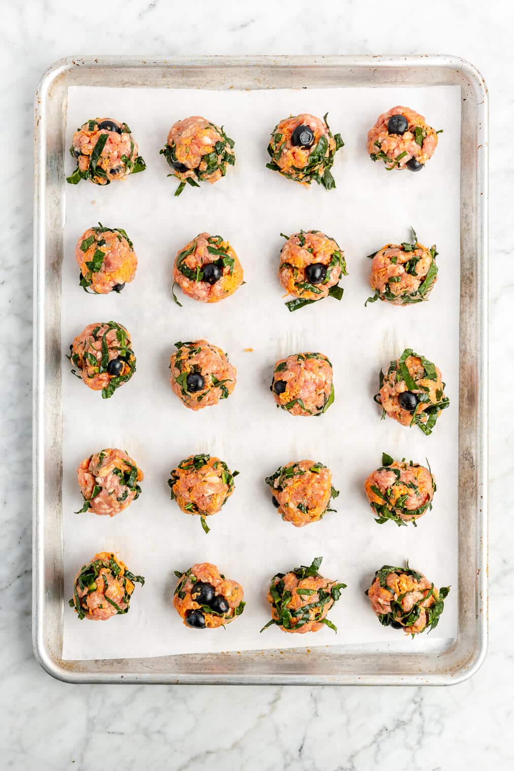 Five rows of 4 blueberry breakfast meatballs on a parchment lined baking sheet on a grey and white marble surface.