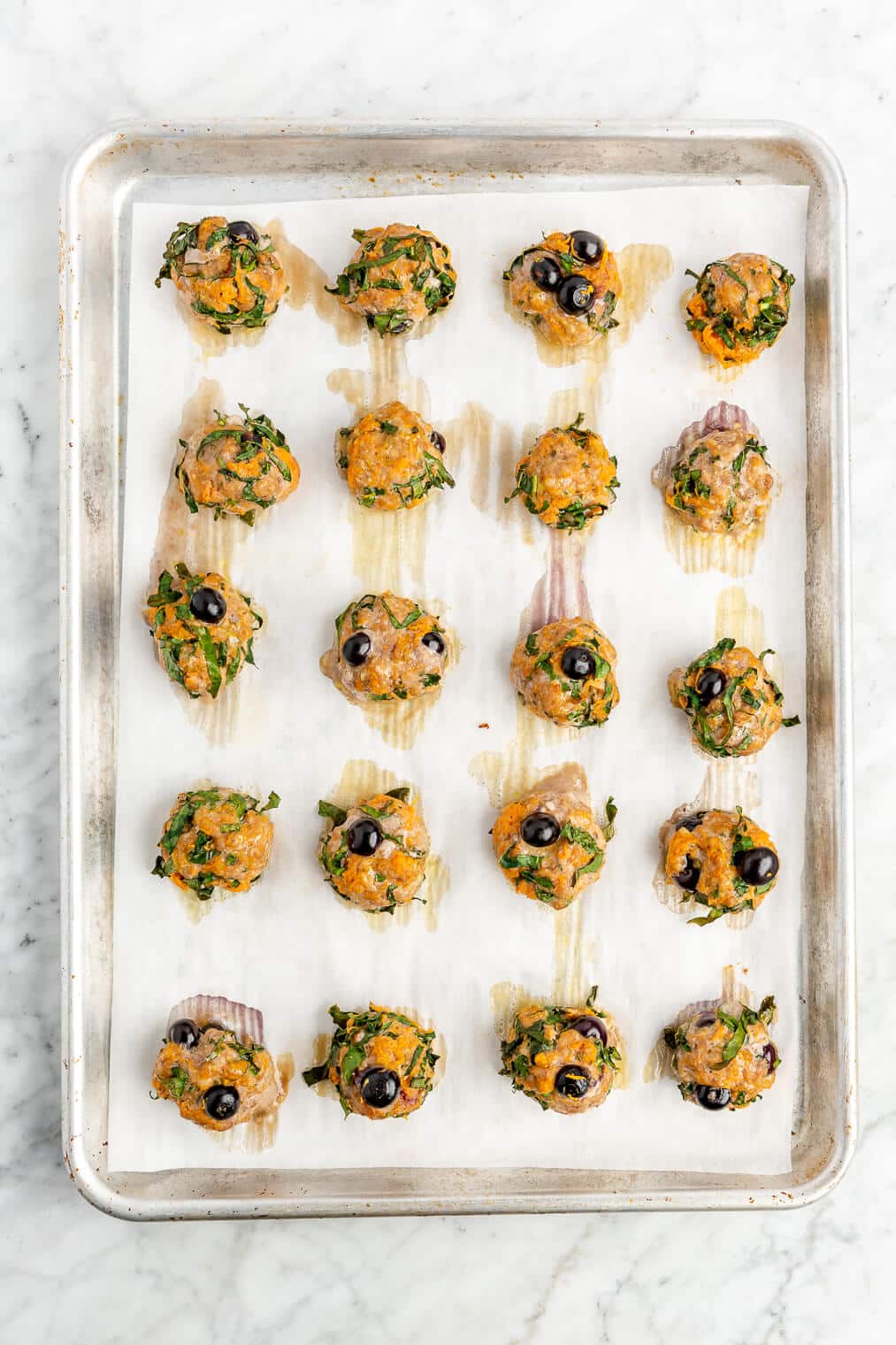 Five rows of four cooked blueberry breakfast meatballs on a parchment lined baking sheet on a grey and white marble surface.