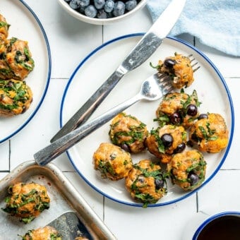 A white plate with a blue rim filled with a serving of breakfast meatballs on a tile surface. There is a fork with a piece of meatball on the fork next to a knife on the plate. There is a white bowl of blueberries and a blue linen to the top and a sheet pan with more blueberries on a metal spatula and a cup of coffee to the bottom.