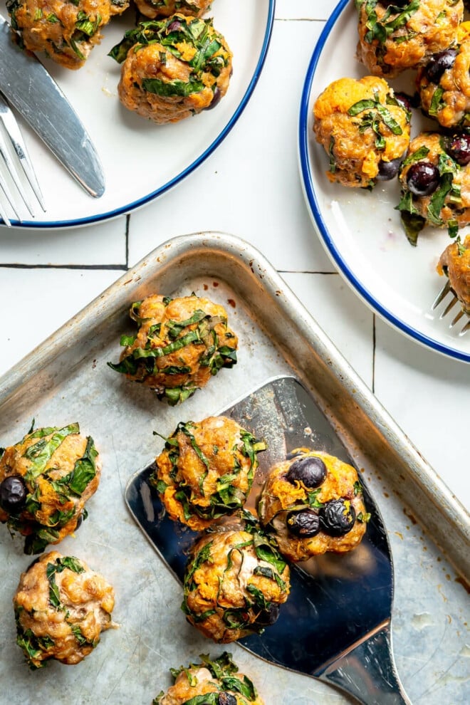 There is a metal spatula with three breakfast meatballs on the spatula on top of a sheet pan with more breakfast meatballs. There are two white plates with blue rims with servings of breakfast meatballs on the plates. All are sitting on a white tile surface.