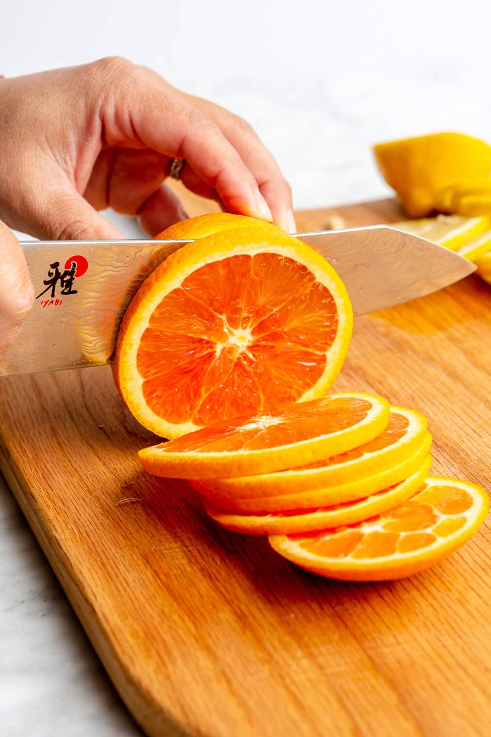 Hand holding an orange and slicing it with a chef's knife on a wooden cutting board.