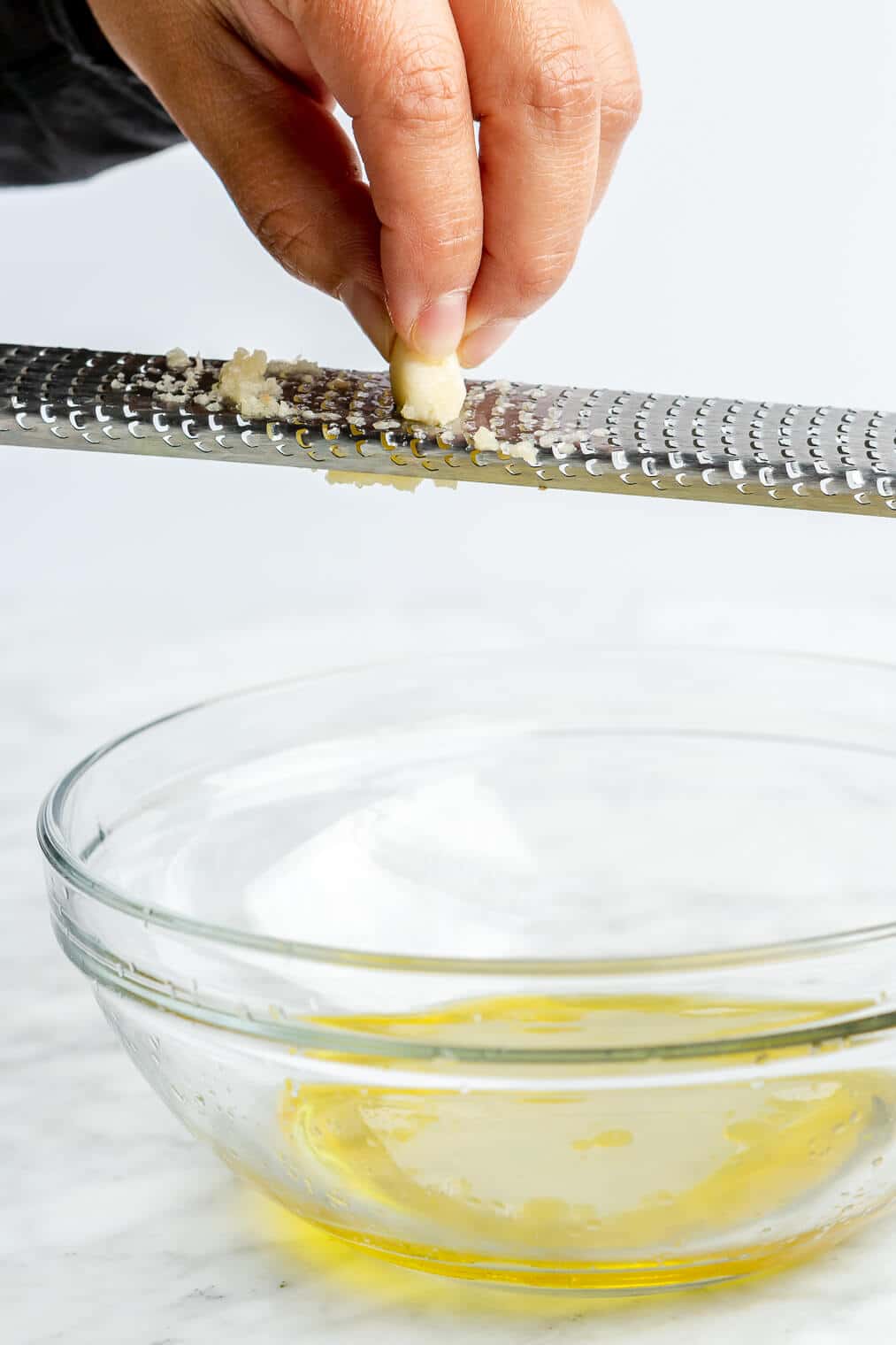 Hand grating a clove of garlic on a microplane into a glass bowl with lemon juice and olive oil.
