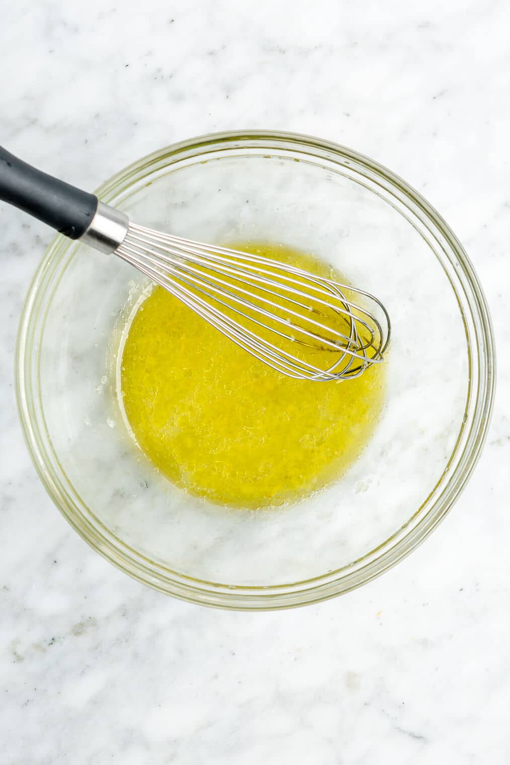 Whisked olive oil, lemon juice, and garlic in a glass bowl on a grey and white marble surface.