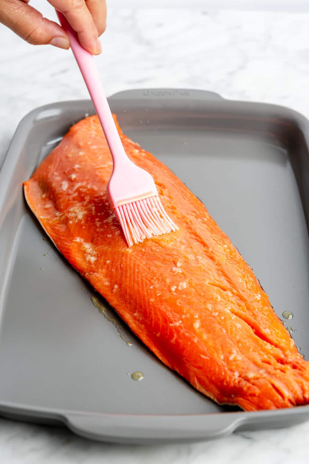 Hand brushing olive oil mixture onto a filet of salmon with a pink silicone brush. The salmon filet is sitting on a grey sheet pan.