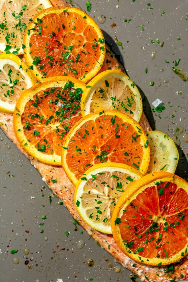 Cooked salmon filet covered with sliced orange and lemon slices sprinkled with parsley on a grey sheet pan.