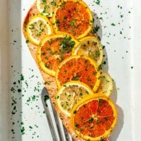 Baked salmon filet covered with sliced orange and lemon slices sprinkled with parsley in a white casserole dish and a metal spatula.