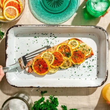 Baked salmon filet covered with sliced orange and lemon slices sprinkled with parsley in a white casserole dish and a metal spatula. The casserole dish is on a linen covered surface alongside two translucent, teal plates, a green glass with ice water, small glass jar with olive oil, parsley, and a bowl of citrus slices. There is a hand holding the metal spatula under the salmon and two hands reading for the water glasses.