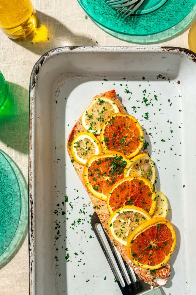Baked salmon filet covered with sliced orange and lemon slices sprinkled with parsley in a white casserole dish and a metal spatula. The casserole dish is on a linen covered surface alongside two translucent, teal plates, a green glass with ice water, and a small glass jar with olive oil.